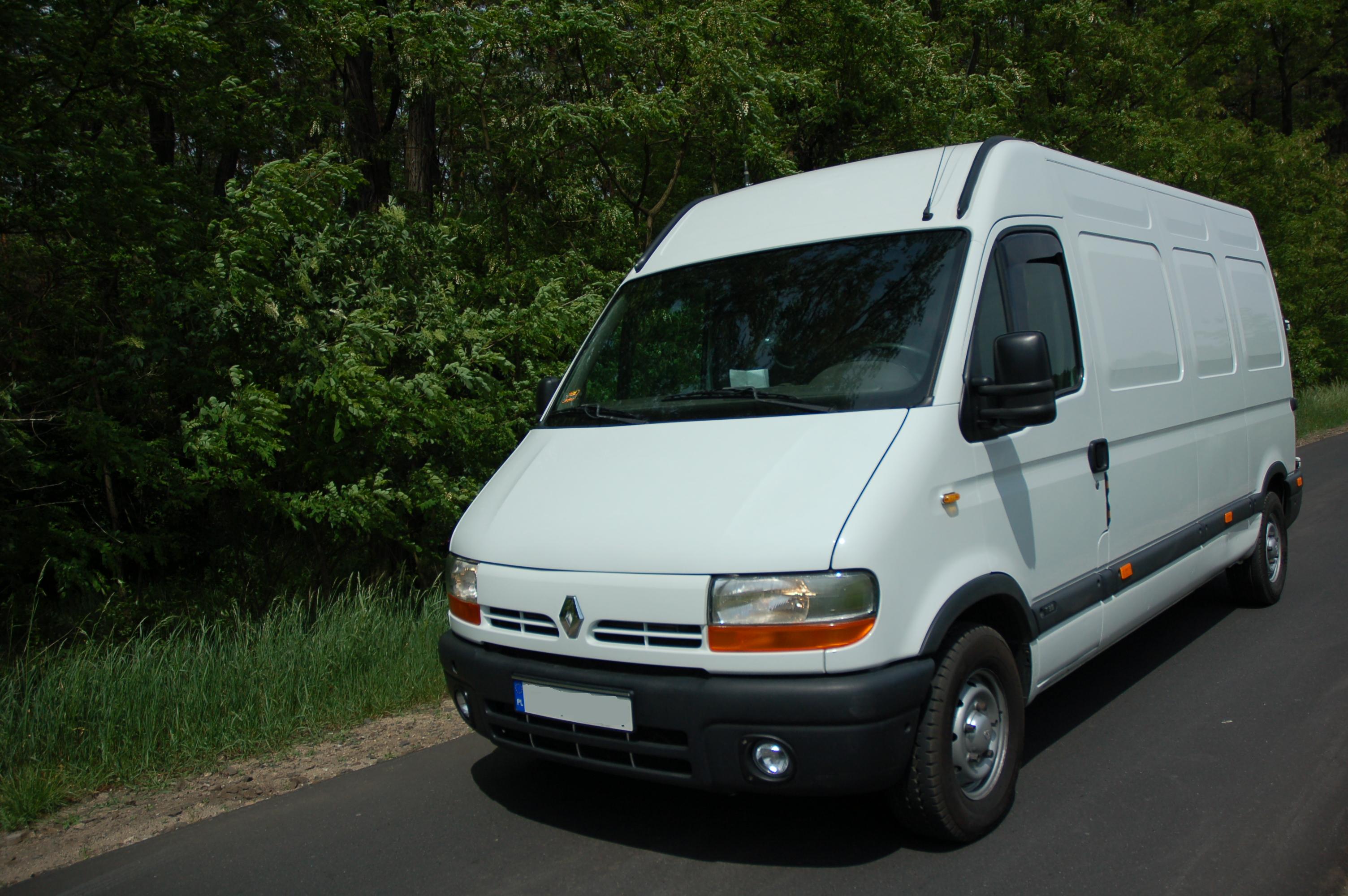 Biały samochód dostawczy Renault Master zaparkowany na asfaltowej drodze, na tle zieleni drzew i trawy, widok z przodu.